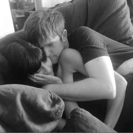 James Blake gives a hug and kiss to Jameela Jamil while sitting in a couch